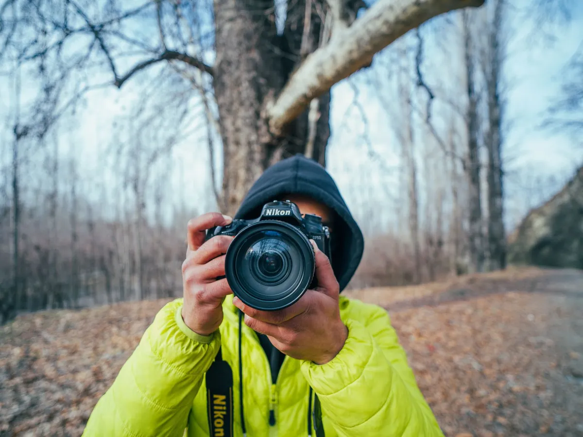 Fotograaf in een gele jas richt een spiegelreflexcamera op de lezer in een herfstachtig bos