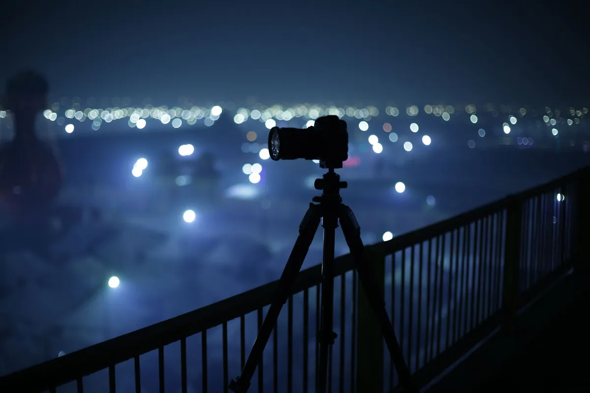 Camera op een statief bij een reling, gericht op de lichtjes van een stad in de avond